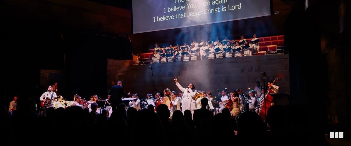 A soloist in white raises her hand during a large-scale worship concert featuring a full orchestra, choir on risers, and projected lyrics reading 'I believe that Jesus Christ is Lord'.
