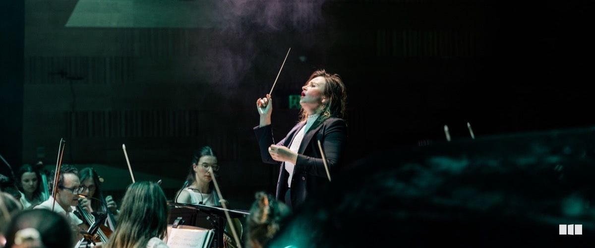 A female conductor raises her baton expressively while leading an orchestra on a dramatically lit stage.