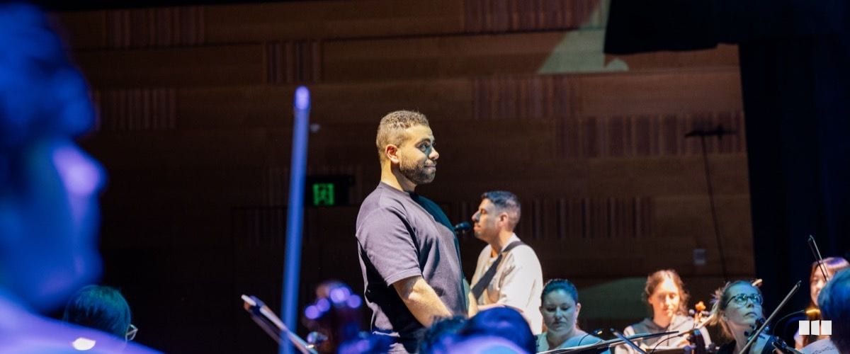 A man in a dark t-shirt stands at centre stage before an orchestra during what appears to be a rehearsal or performance in a concert hall.