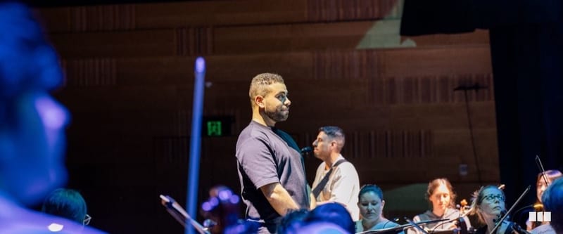 A man in a dark t-shirt stands at centre stage before an orchestra during what appears to be a rehearsal or performance in a concert hall.