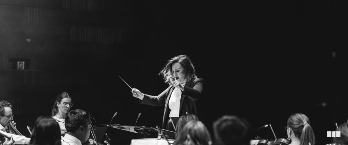 A female conductor leads an orchestra with passionate intensity, baton raised and mouth open mid-direction.