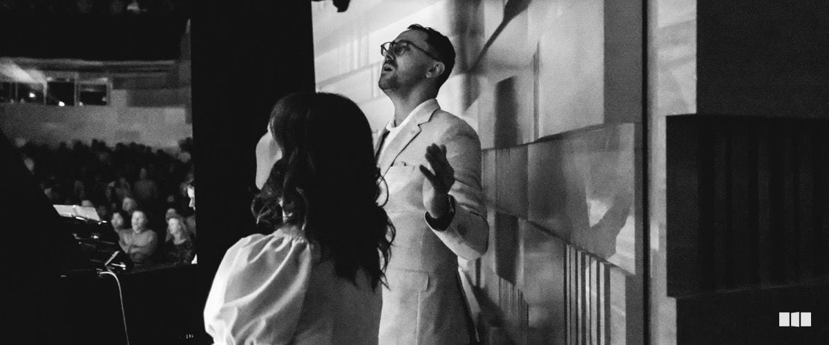 A male soloist in a white suit gazes upward while performing on stage alongside a female singer before a seated audience.