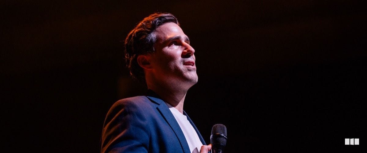 A male soloist in a navy suit looks upward while holding a microphone on a darkened stage under warm spotlight