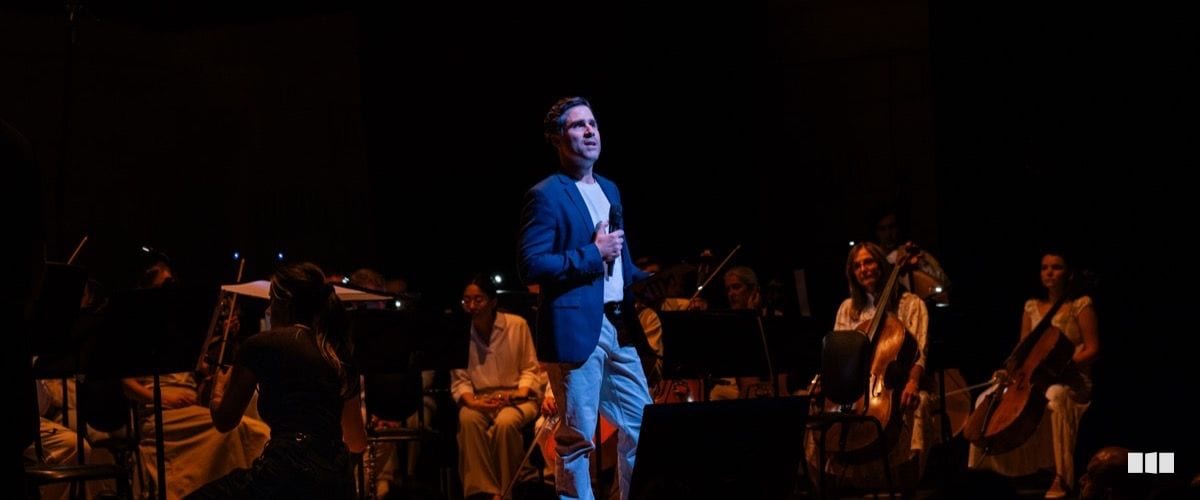 A male soloist in a blue blazer holds a microphone and sings centre-stage while orchestra musicians are seated behind him in a darkened concert hall.