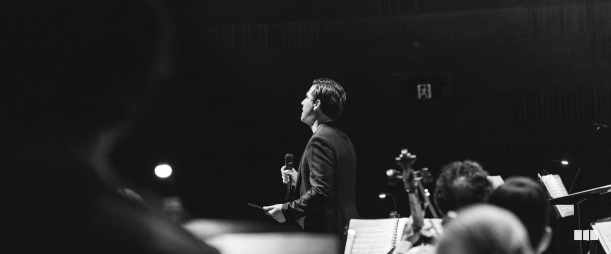 A man in a dark suit speaks into a microphone on stage in front of an orchestra during a concert.