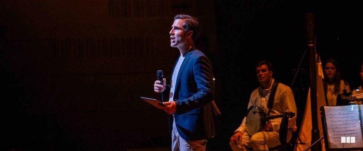 A male MC or presenter speaks into a handheld microphone on a darkly lit stage while musicians sit in the background