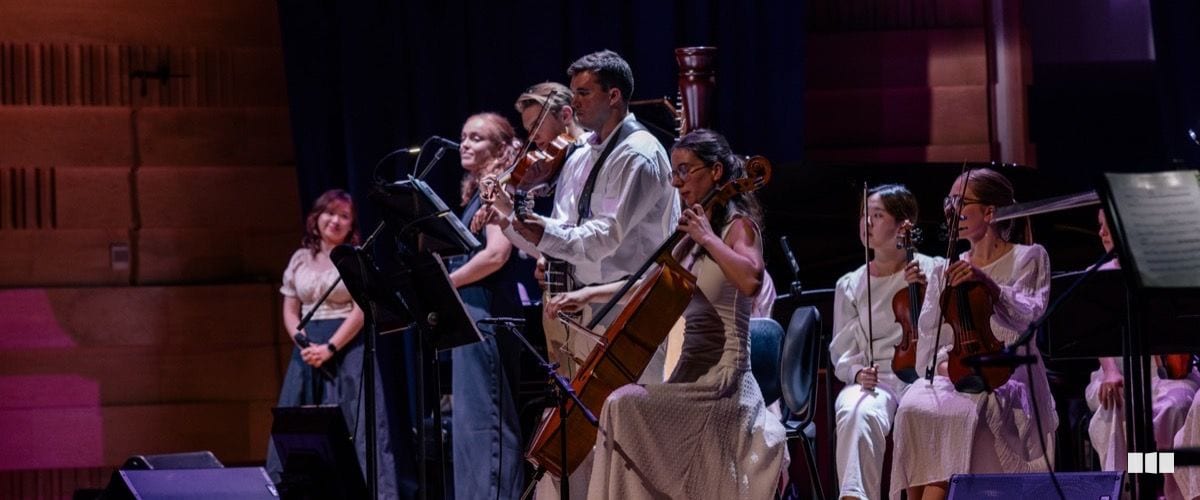 Chamber orchestra musicians perform on stage, with string players and a harpist visible under warm concert lighting.
