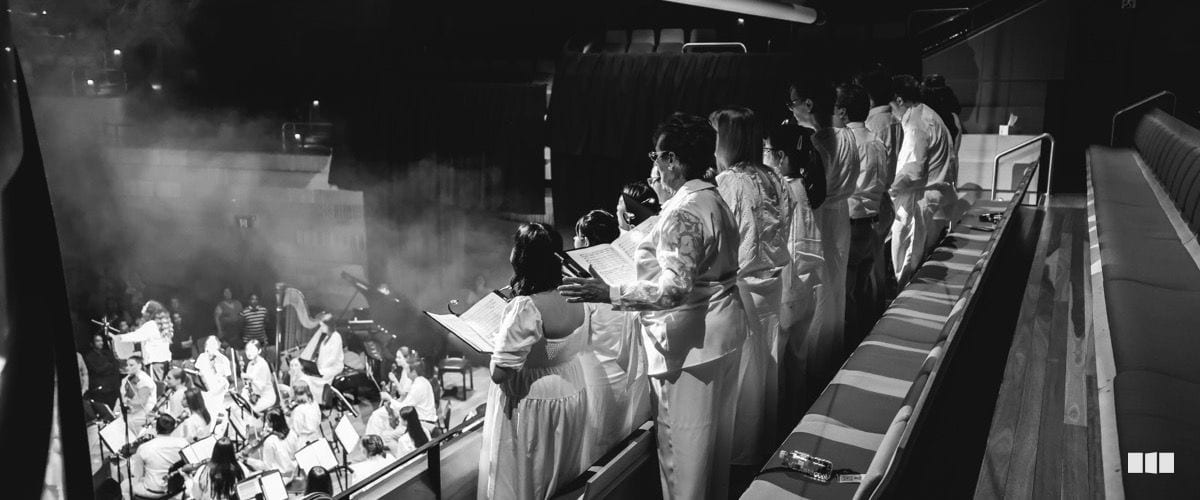 A choir in white robes sings from sheet music on a balcony above a full orchestra performing on stage in a dramatic, smoke-lit venue.