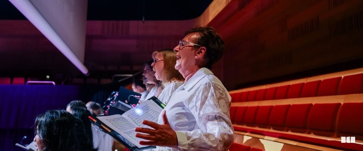 A row of choir singers in white shirts perform on stage in a concert hall, holding sheet music folders under dramatic lighting