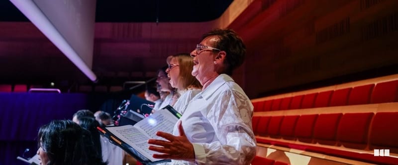 A row of choir singers in white shirts perform on stage in a concert hall, holding sheet music folders under dramatic lighting