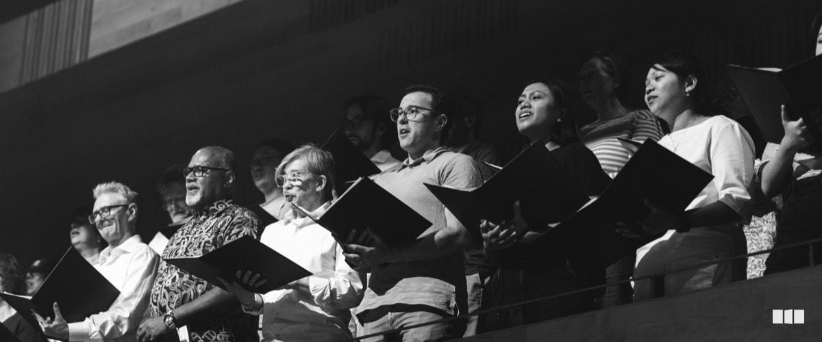 A diverse choir ensemble sings from sheet music folders during a performance or rehearsal