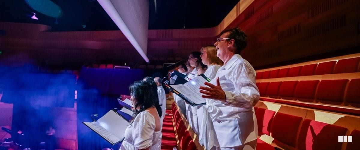 Choir members dressed in white sing from illuminated music folders in the aisle of a concert hall with red seating