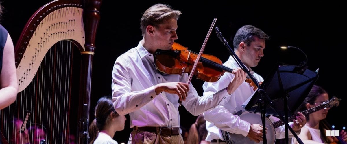 Two violinists in white shirts perform on stage with a harp visible in the background during an orchestral concert