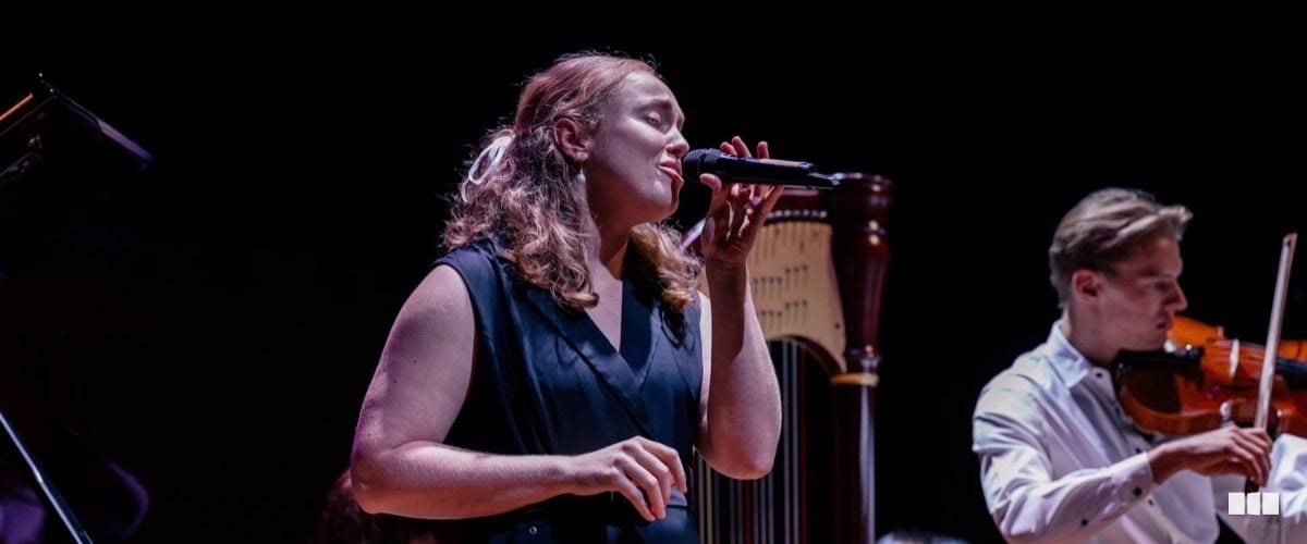 A female soloist sings into a handheld microphone on stage while a violinist performs in the background beside a harp.