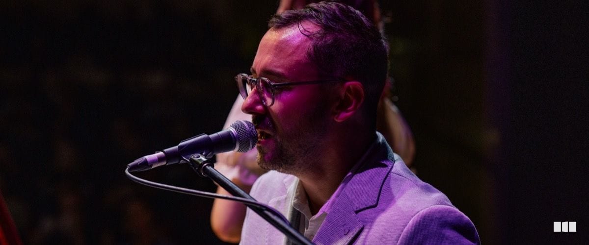 A man wearing glasses and a light purple blazer sings into a microphone under purple stage lighting.