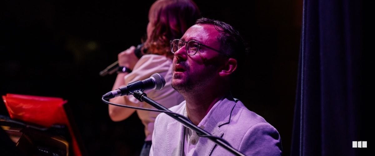 A man wearing glasses and a light blazer sings into a microphone on a darkened stage, with a second performer visible in the background