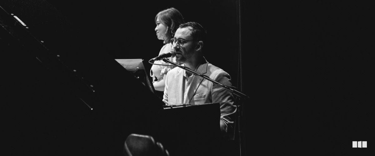A man wearing glasses and a light suit jacket sings into a microphone at a keyboard, with a woman visible behind him on a darkened stage.