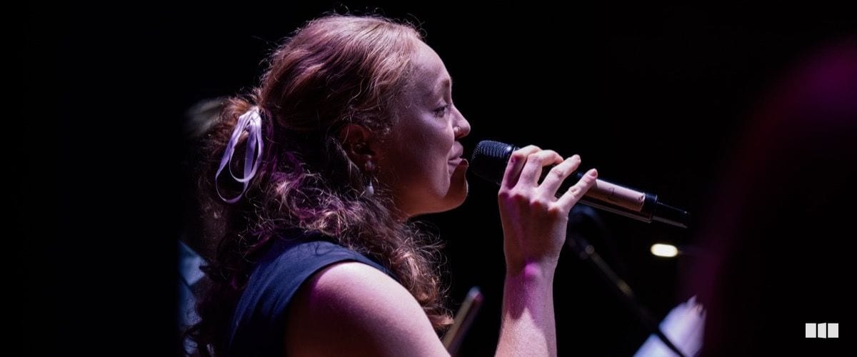 A female vocalist performs on stage, singing into a handheld microphone under warm stage lighting.