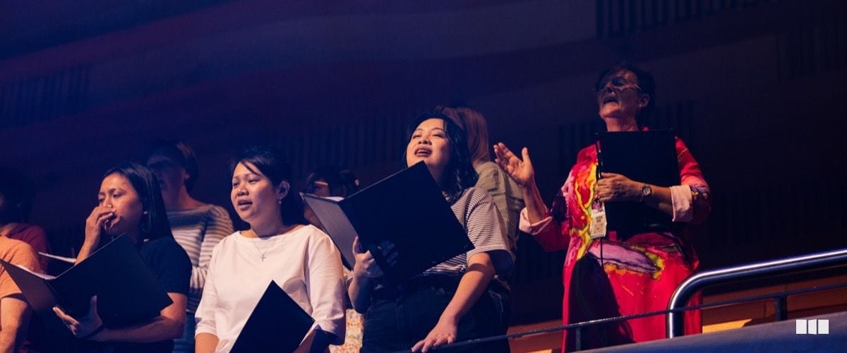 Several women holding black folders sing with expressive faces during what appears to be a choral performance in a large auditorium.