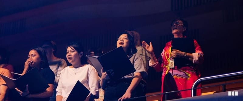Several women holding black folders sing with expressive faces during what appears to be a choral performance in a large auditorium.
