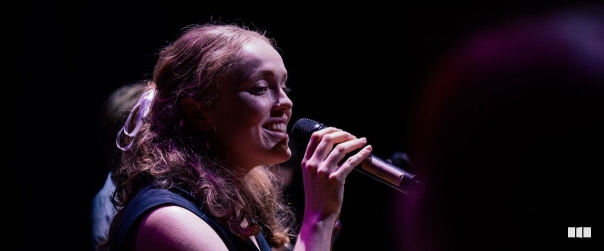 A young woman with curly auburn hair smiles as she sings into a handheld microphone under stage lighting