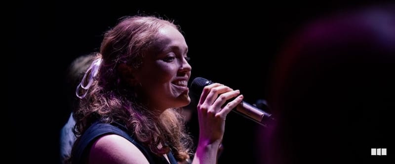 A young woman with curly auburn hair smiles as she sings into a handheld microphone under stage lighting