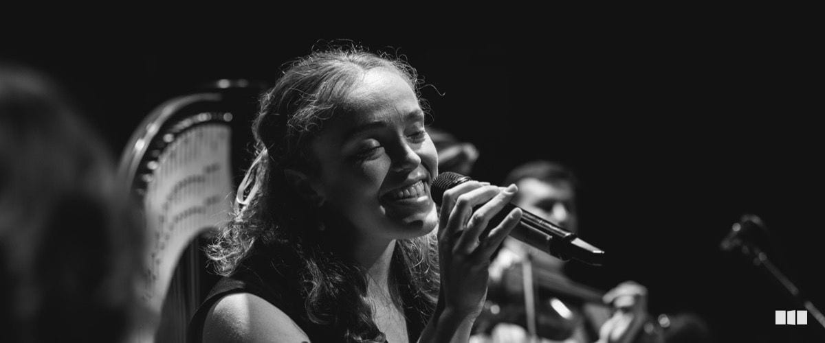 A young woman smiles warmly as she sings into a handheld microphone, with orchestral musicians visible in the background.