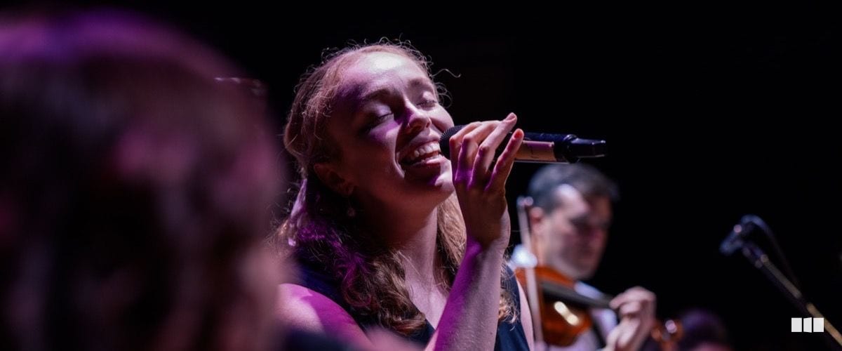 A female soloist sings with eyes closed and a broad smile into a handheld microphone, lit by warm purple stage lighting, with a violinist visible in the background.