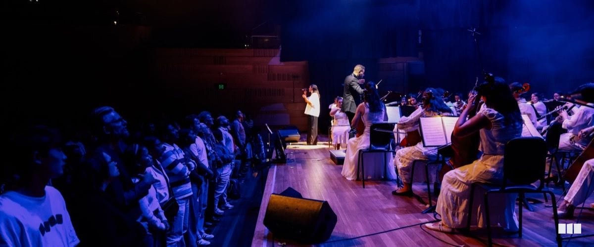A conductor leads a soloist and full orchestra with string players in white attire on a warmly lit concert stage before a standing audience bathed in blue light