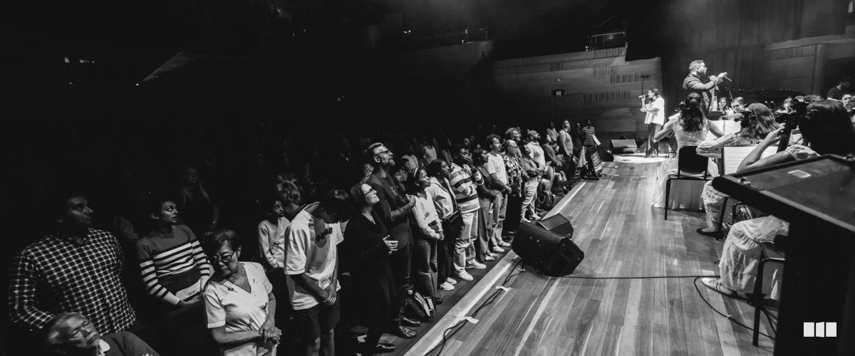A large crowd of audience members stands at the front of the stage during a live indoor concert performance