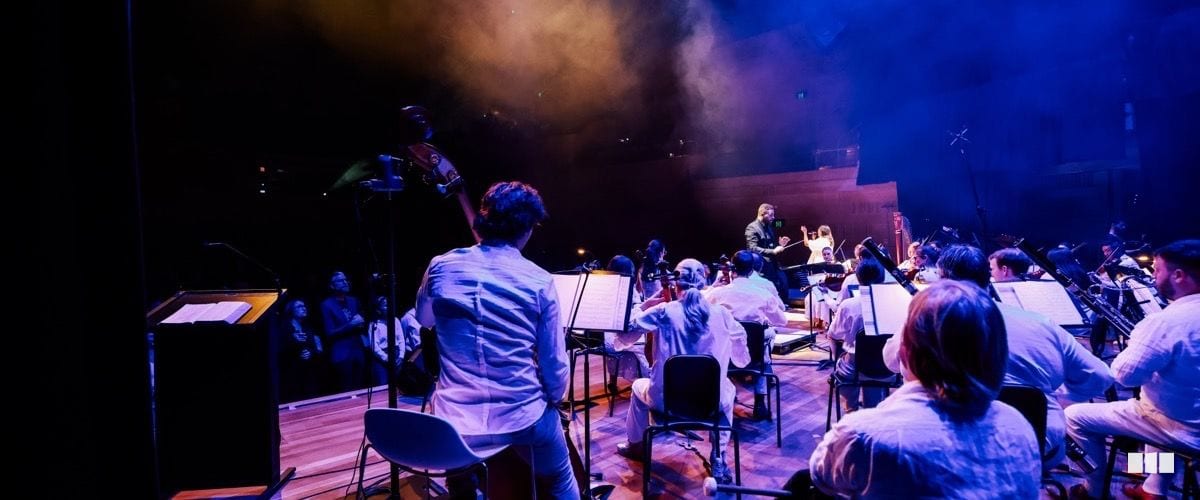 An orchestra dressed in white performs on a dramatically lit stage under blue and purple atmospheric lighting, viewed from behind a musician toward the conductor.