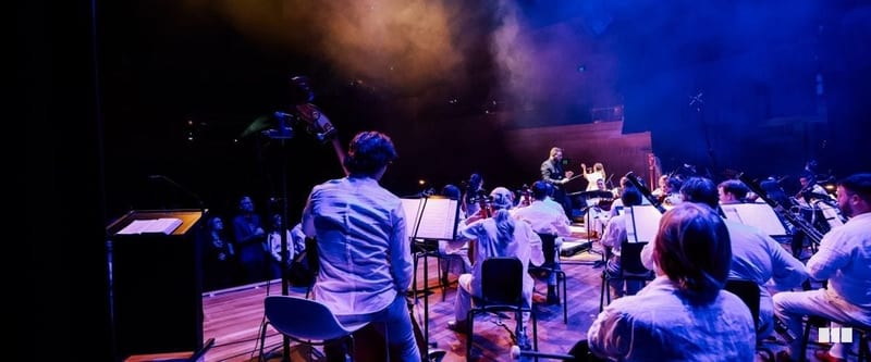 An orchestra dressed in white performs on a dramatically lit stage under blue and purple atmospheric lighting, viewed from behind a musician toward the conductor.