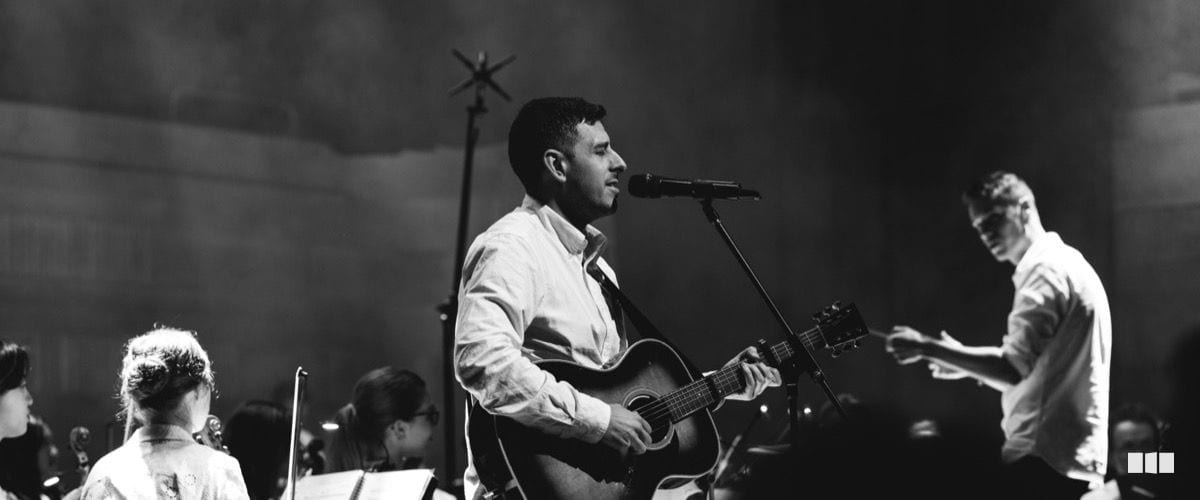 A male guitarist and vocalist performs at a microphone centre-stage with an orchestra ensemble visible behind him and a conductor gesturing to the right.