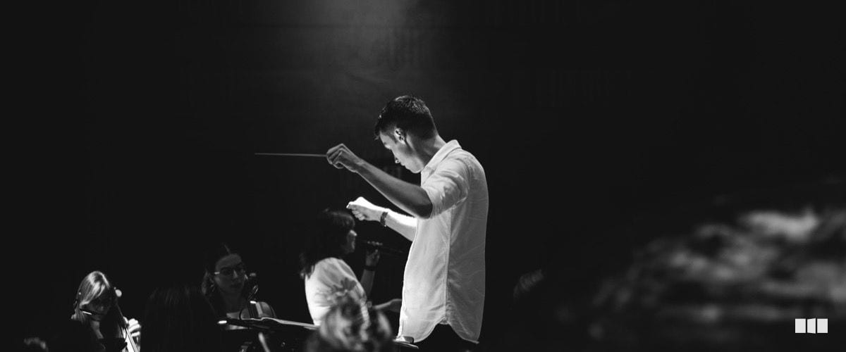 A conductor leads an orchestra from the podium, baton raised, illuminated by a dramatic spotlight in a darkened concert hall.