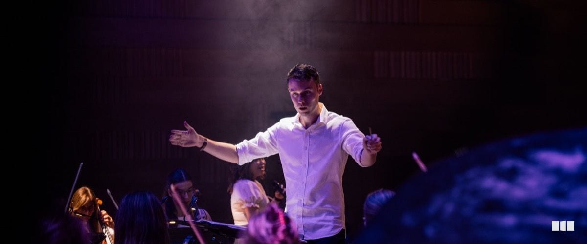 A conductor in a white shirt leads the orchestra with arms outstretched under dramatic stage lighting.
