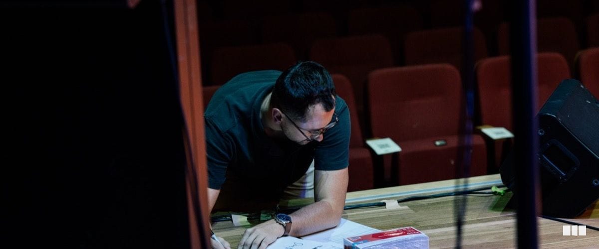 A man wearing glasses and a dark t-shirt leans over a table reviewing documents or scores in an empty theatre auditorium