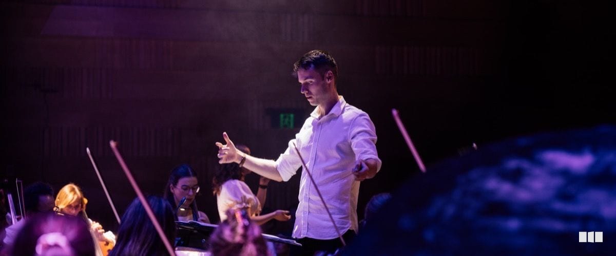 A conductor in a white shirt leads the orchestra with baton in hand under dramatic stage lighting.