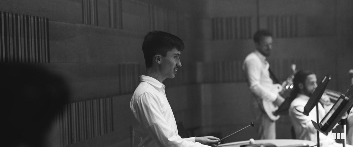 A young musician plays percussion at a keyboard instrument during an orchestra rehearsal in an acoustic studio, with other musicians visible in the background.
