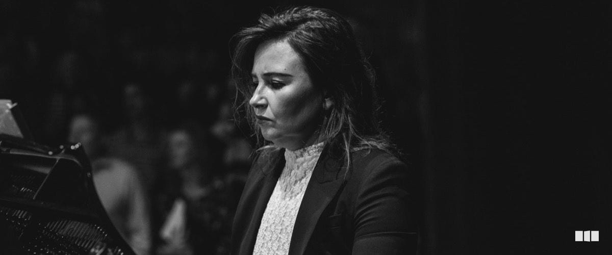 A woman in a dark blazer and lace top plays piano with focused concentration during an orchestral performance.