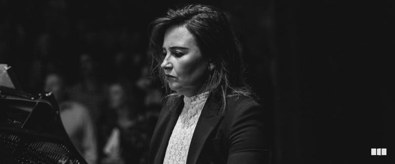 A woman in a dark blazer and lace top plays piano with focused concentration during an orchestral performance.