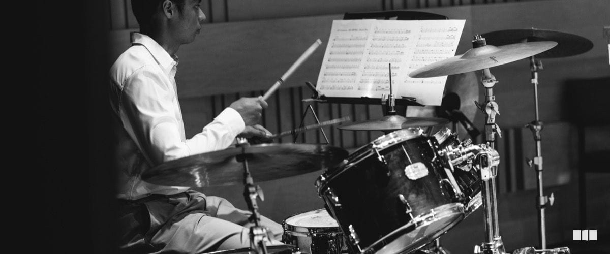A drummer plays at a kit during an orchestral performance, reading from sheet music on a stand.