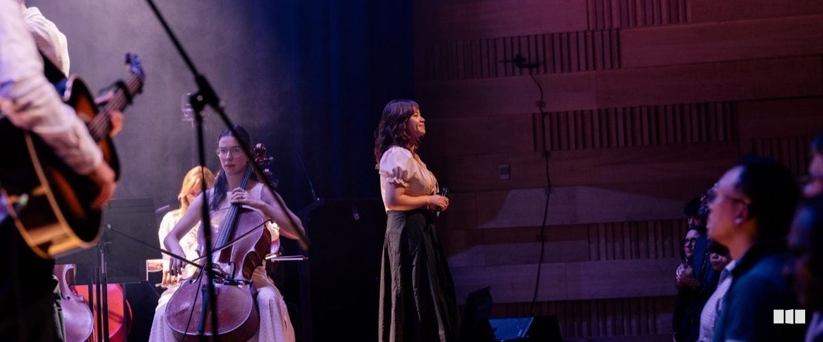 A cellist performs on stage while a vocalist stands centre-stage during an orchestral concert in a warmly lit concert hall
