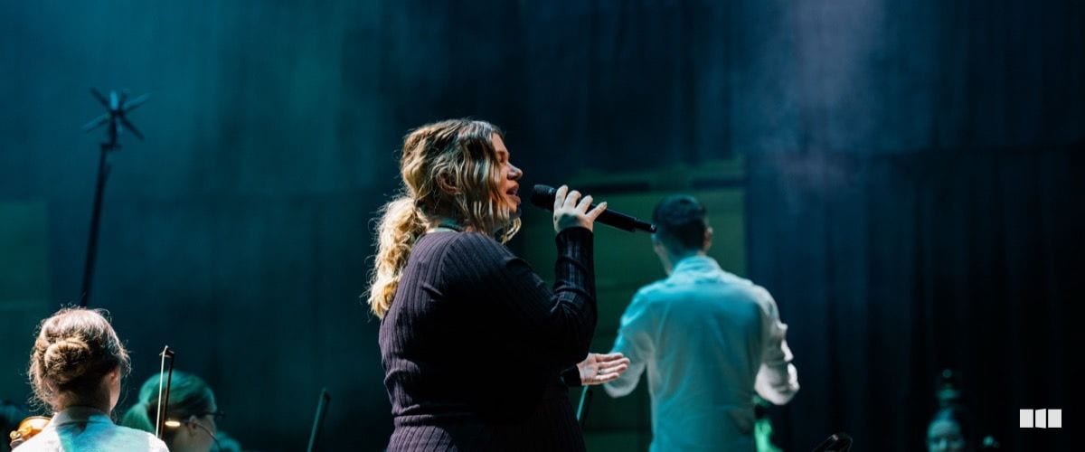 A female vocalist performs on stage with a handheld microphone, backed by orchestra musicians in atmospheric blue stage lighting