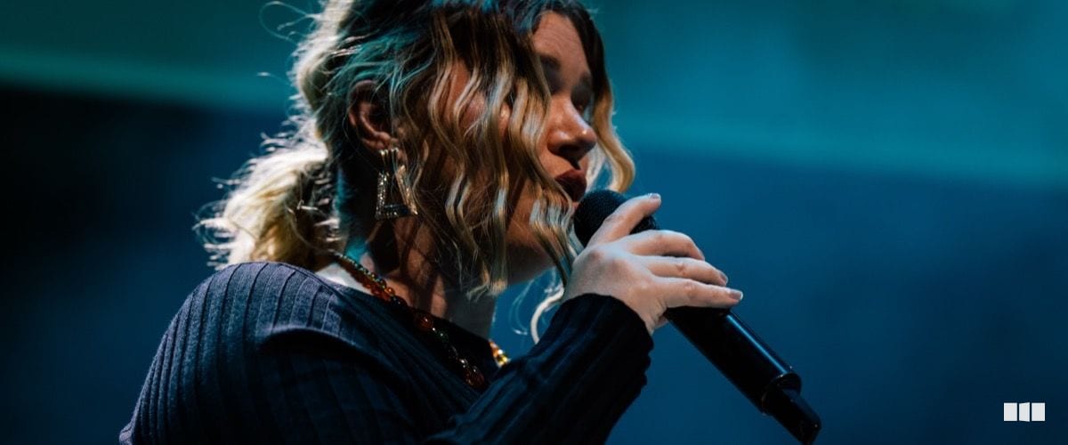 A female vocalist performs on stage, holding a microphone close to her lips under teal-toned stage lighting.