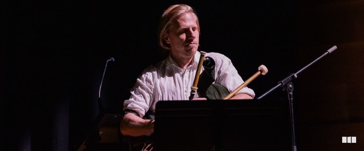 A blonde male musician sits at a keyboard instrument with a mallet percussion instrument visible beside him on a darkened stage