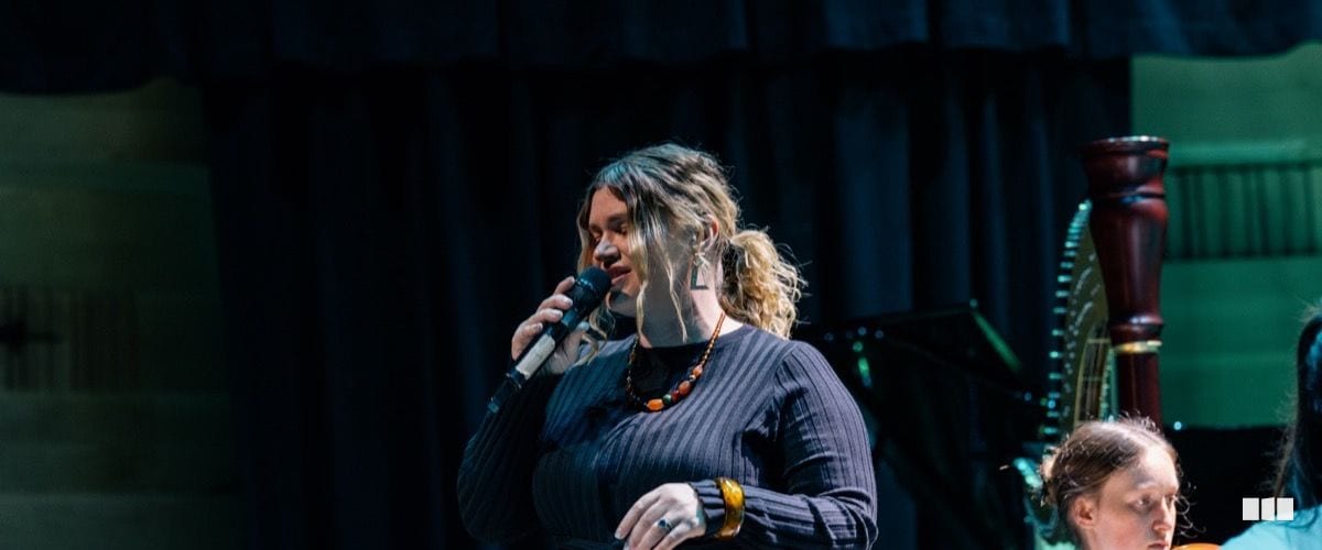 A female soloist sings into a handheld microphone on a darkened concert stage, with a harp visible in the background.