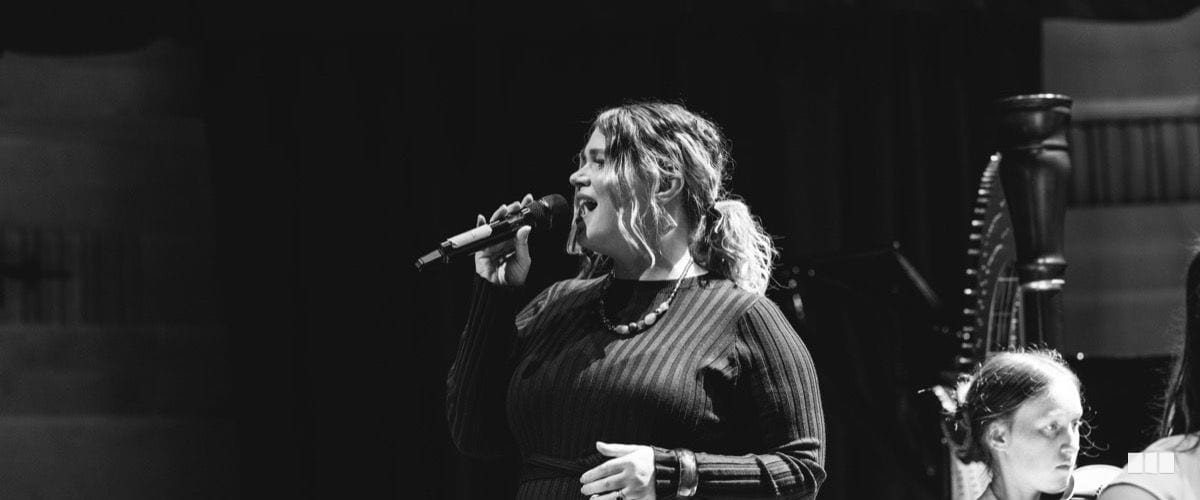 A female soloist sings passionately into a handheld microphone on stage, with a harpist visible in the background.