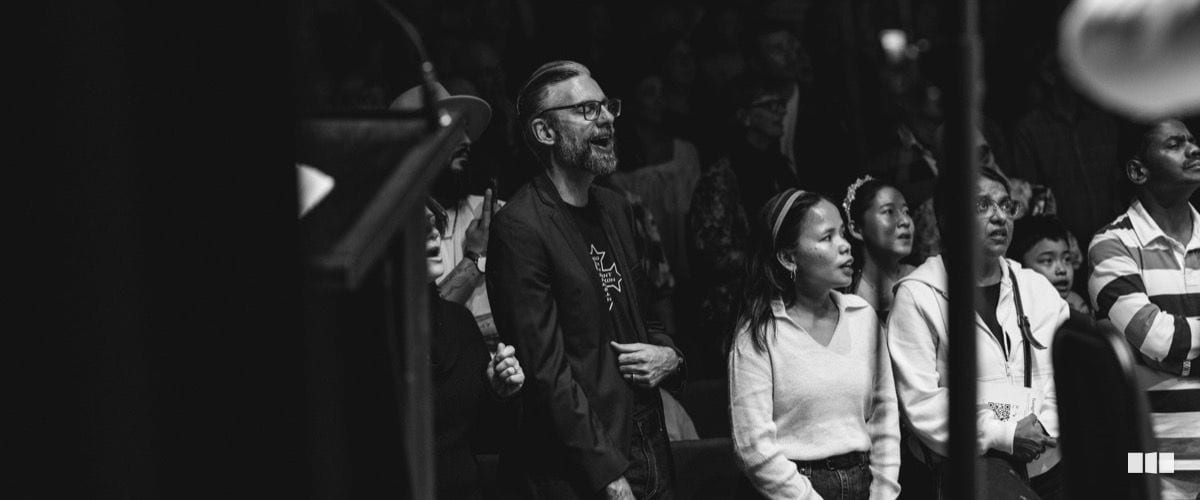 A bearded man wearing glasses and a dark blazer sings expressively alongside a diverse group of audience members or choir participants during a concert event.