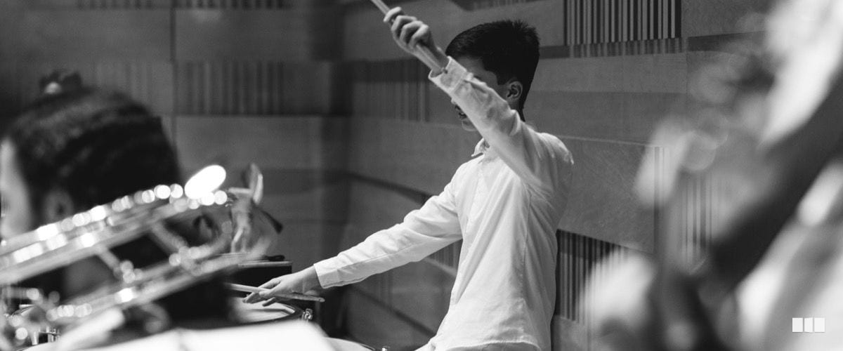 A young drummer plays with expressive energy, one stick raised high, during an orchestra rehearsal or performance.