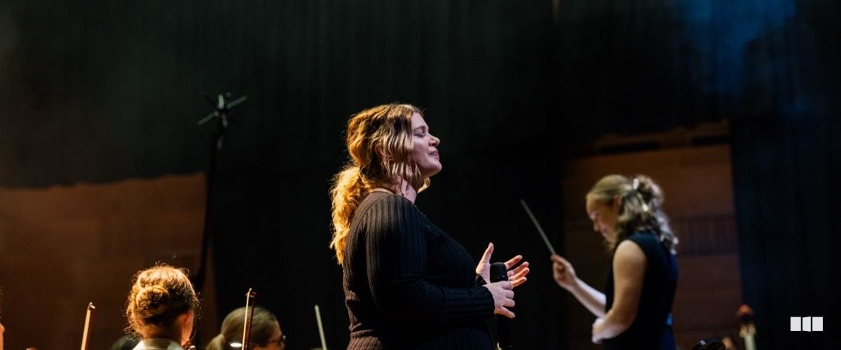 A soprano soloist performs with eyes closed and hands clasped while a conductor leads the orchestra behind her on a darkened concert stage.